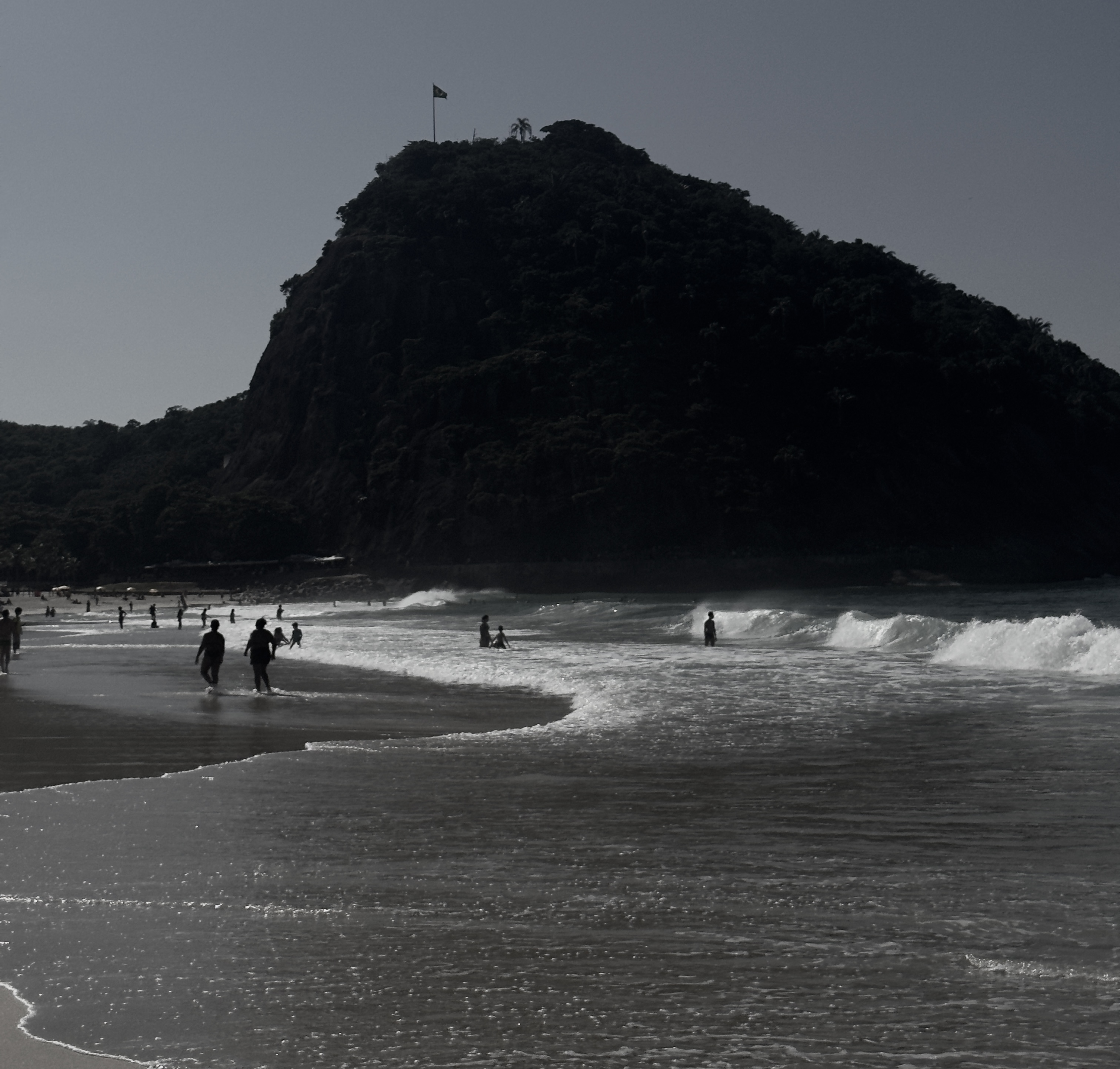 Beachgoers and breaking waves along the shoreline beneath a tree-covered hill
